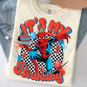 May include: A cream-colored t-shirt featuring a graphic of a red and blue superhero wearing a party hat, holding a birthday cake with candles. The design includes a black and white checkered circle, red lightning bolts, balloons, and the text IT'S MY Birthday.