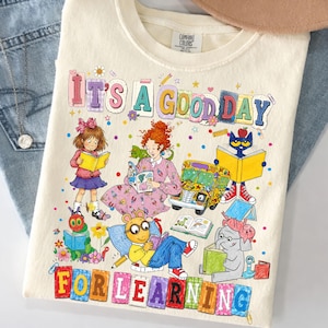 May include: A cream-colored t-shirt featuring a colorful graphic with the text "IT'S A GOOD DAY FOR LEARNING." The illustration includes various children's book characters, a school bus, and books, surrounded by small confetti-like dots and flowers.