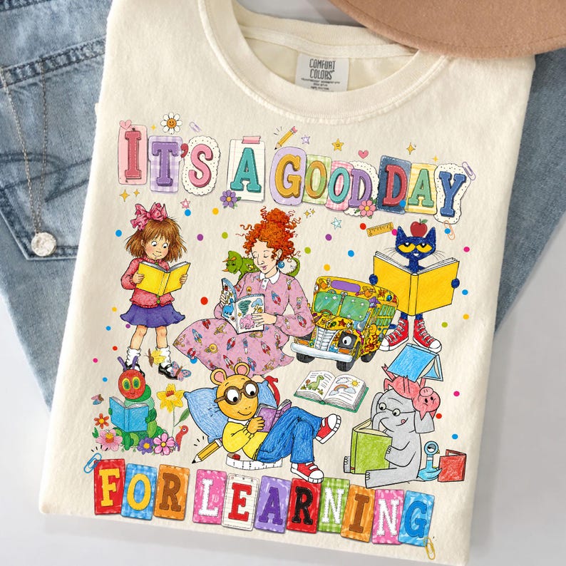 Puede incluir: Una camiseta de color crema con un gr&aacute;fico colorido y el texto "IT'S A GOOD DAY FOR LEARNING". La ilustraci&oacute;n incluye varios personajes de libros infantiles, un autob&uacute;s escolar y libros, rodeados de peque&ntilde;os puntos y flores.