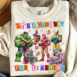 May include: A cream-colored crewneck sweatshirt featuring a graphic of Marvel superheroes reading books. The text reads IT'S A GOOD DAY FOR LEARNING. Characters include Hulk, Spider-Man, Iron Man, Wolverine, Thor, and Captain America with small flowers.