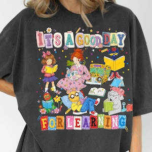 Puede incluir: Una camiseta gris carb&oacute;n con un estampado gr&aacute;fico colorido que incluye el texto IT'S A GOOD DAY FOR LEARNING. El dise&ntilde;o muestra ilustraciones de varios personajes de libros infantiles leyendo, un autob&uacute;s escolar amarillo y confeti.