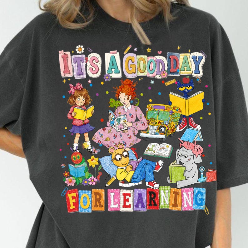 Puede incluir: Una camiseta gris carb&oacute;n con un estampado gr&aacute;fico colorido que incluye el texto IT'S A GOOD DAY FOR LEARNING. El dise&ntilde;o muestra ilustraciones de varios personajes de libros infantiles leyendo, un autob&uacute;s escolar amarillo y confeti.