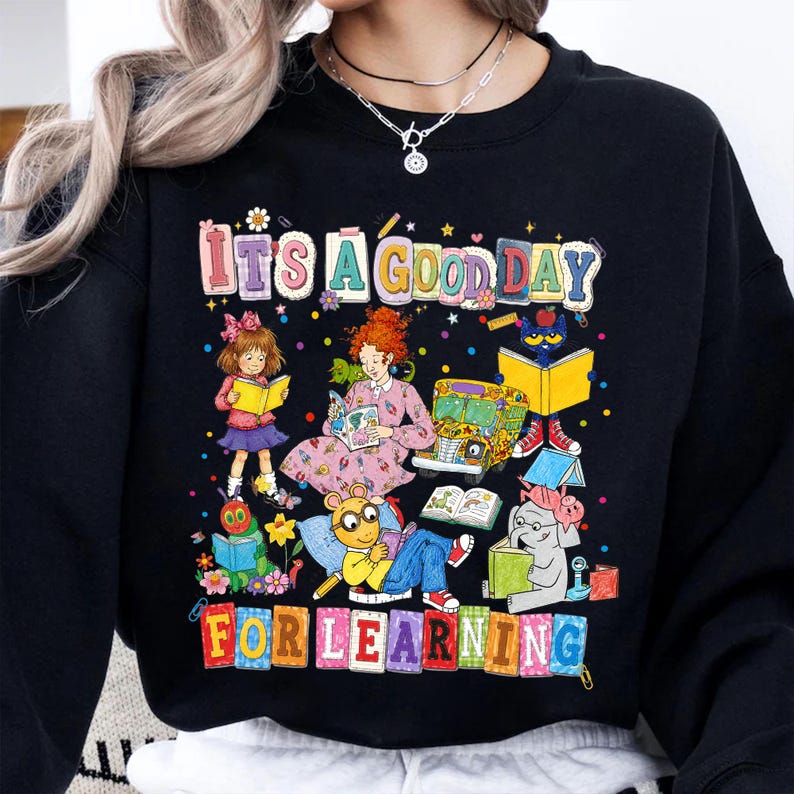 Puede incluir: Una sudadera negra de cuello redondo con un estampado gr&aacute;fico colorido con el texto "IT'S A GOOD DAY FOR LEARNING". El dise&ntilde;o incluye ilustraciones de varios personajes de libros infantiles leyendo, un autob&uacute;s escolar y confeti disperso.