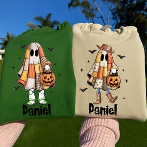 May include: A dark green sweatshirt and a beige sweatshirt, each featuring a graphic of a ghost wearing a patchwork blanket, holding a jack-o'-lantern, and surrounded by bats and stars. The name Daniel is printed in black below each ghost.