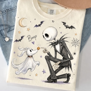 Puede incluir: Una camiseta de color crema con una ilustración gráfica de un personaje esqueleto con traje de rayas arrodillado para tocar la nariz de un perro fantasma con una pequeña calabaza. El diseño incluye murciélagos, estrellas, una luna y el texto Boop.