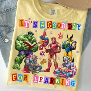May include: A pale yellow t-shirt featuring a graphic print of Marvel superheroes including Hulk, Spider-Man, Iron Man, Wolverine, Thor, and Captain America reading books. The text reads IT'S A GOOD DAY FOR LEARNING in colorful block letters.