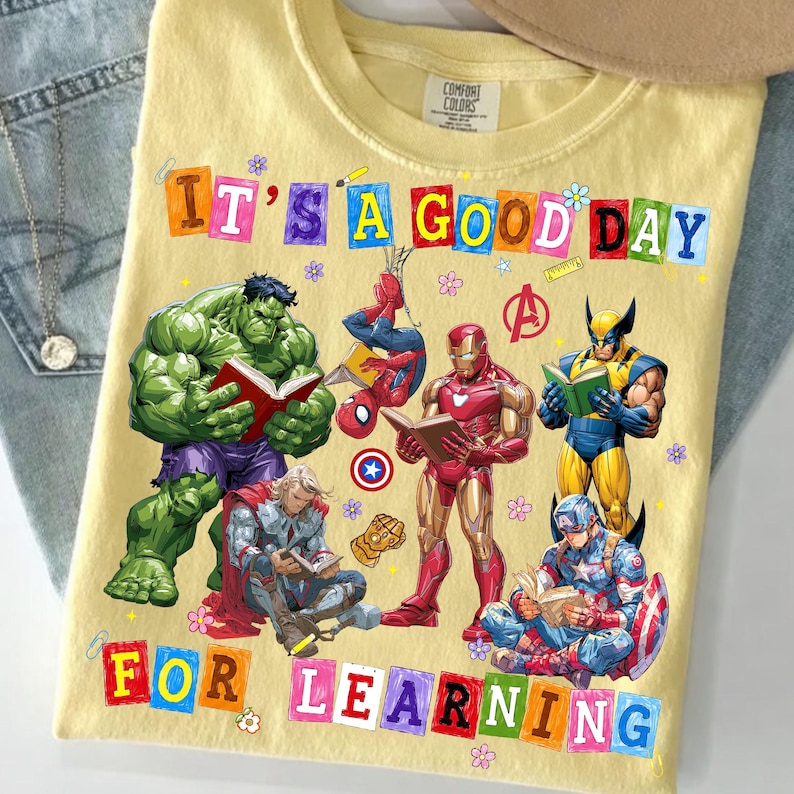 May include: A pale yellow t-shirt featuring a graphic print of Marvel superheroes including Hulk, Spider-Man, Iron Man, Wolverine, Thor, and Captain America reading books. The text reads IT'S A GOOD DAY FOR LEARNING in colorful block letters.