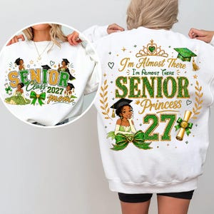 May include: A white sweatshirt featuring a graphic print of a princess in a graduation cap and gown. The text reads I'm Almost There Senior Princess 27, accompanied by a gold crown, a diploma, and green glitter accents. The design includes floral motifs.