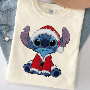 May include: A cream-coloured t-shirt featuring an embroidered graphic of a blue alien character wearing a red and white Santa Claus suit and hat. The shirt has a Comfort Colors brand label at the inner neckline. The design is centred on the front.
