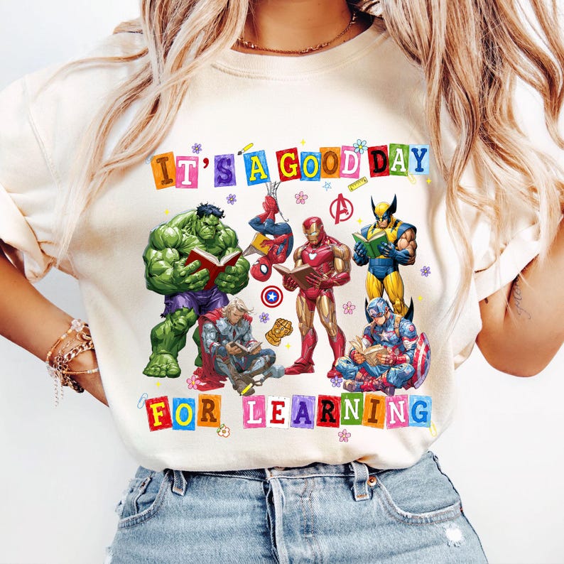 May include: A cream-colored t-shirt featuring a graphic of Marvel superheroes including Hulk, Spider-Man, Iron Man, Wolverine, Thor, and Captain America reading books. The text reads IT'S A GOOD DAY FOR LEARNING in colorful block letters.