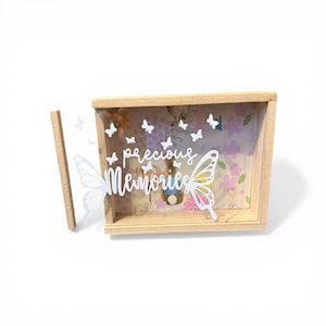 Peut inclure: Une boîte à souvenirs en bois avec un fond floral et l'inscription "precious memories" en écriture blanche. Des découpes de papillons blancs sont éparpillées autour de la boîte, avec un grand papillon sur le devant.