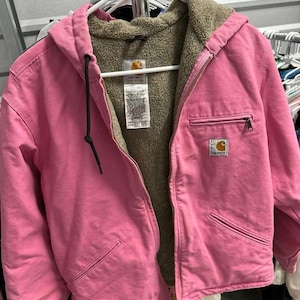May include: A pink Carhartt hooded jacket with a brown sherpa lining. The jacket has a zipper closure, multiple pockets, and the Carhartt logo patch. The jacket is hanging on a white hanger.