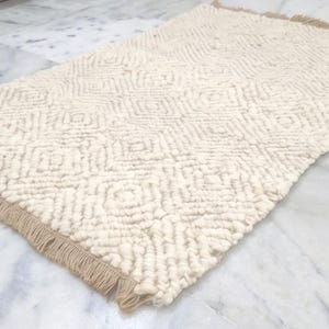 May include: Cream-colored rectangular rug with a geometric diamond pattern. The rug has a textured, woven appearance and a tan fringe along one edge. The rug is made of a soft material and is suitable for a living room or bedroom.