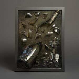 May include: A framed artwork featuring a shattered champagne bottle. The dark glass fragments are arranged against a textured background within a black frame. The bottle's label is visible.