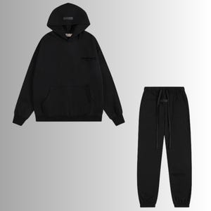 Op de afbeelding: Zwarte hoodie en joggingbroek set. De hoodie heeft een capuchon, een voorzak en het woord "ESSENTIALS" op de borst gedrukt. De joggingbroek heeft een taille met trekkoord en het woord "ESSENTIALS" op de broekspijp. De set is gemaakt van zacht materiaal.