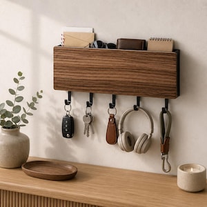 May include: A wall-mounted rectangular wooden organizer with a dark wood grain finish featuring a top storage slot and five black metal hooks. The hooks hold a car key fob, a set of keys, a leather keychain, wireless headphones, and a strap.