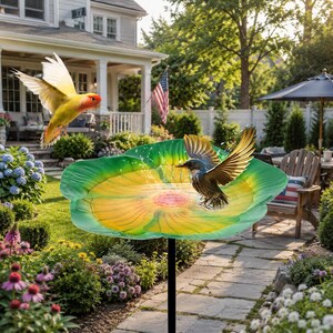 Könnte beinhalten: Ein dekoratives Vogelbad mit einem grün-gelben Blumendesign, das von einer schwarzen Metallsäule getragen wird. Zwei Vögel sind in der Nähe des Vogelbades zu sehen, mit spritzendem Wasser. Das Vogelbad befindet sich in einem Garten.
