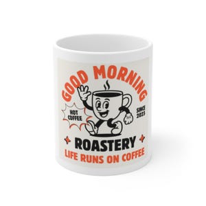 Op de afbeelding: Witte keramische koffiemok met een retro design. De mok heeft de woorden "GOOD MORNING" in oranje, samen met een cartoon koffiekopje karakter. Extra tekst bevat "HOT COFFEE", "ROASTERY" en "LIFE RUNS ON COFFEE".