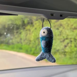Op de afbeelding: Een naaldgevilte vis-ornament in blauw- en grijstinten, voorzien van een opvallend wit oog met een zwarte pupil. De vis heeft een textuurrijk, pluizig oppervlak en hangt aan een dun zwart koord, bedoeld om aan een achteruitkijkspiegel te hangen.