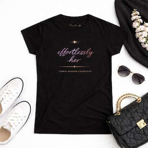 May include: Black t-shirt with the text "effortlessly her" in a gradient pink and purple font. The shirt also features the text "CHERRY BLOSSOM COLLECTION". The image includes white sneakers, a black handbag, sunglasses, and a black hat.