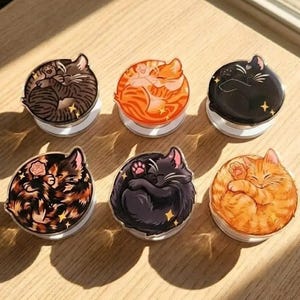 May include: Six round phone grips featuring sleeping cat designs. The cats are depicted in various colors, including brown, orange, and black, with star accents. Each grip has a white base.
