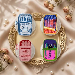 Peut inclure: Quatre accessoires en forme de boîtes de sardines colorées. Les motifs comprennent une boîte bleue avec le mot "Sardines", une boîte avec des poissons rouges et oranges, une boîte rose avec "Brisling Sardines", et une boîte avec un chat noir et des poissons.