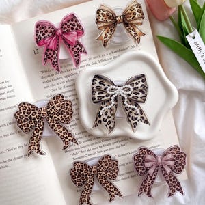 May include: Several bow-shaped phone grips with a leopard print design. Some have pink or brown details, and one is adorned with pearls. They are arranged on a book and a white dish, offering a stylish accessory.