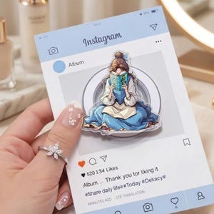 May include: A light blue Instagram-themed notepad with a sticker of a woman in a blue dress reading a book. The notepad has the Instagram logo and the text "Album... Thank you for liking it." A silver ring with a flower design is also visible.