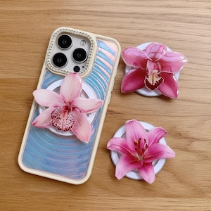 May include: A phone case with a floral design and three matching flower phone grips. The phone case has a cream-colored border and a blue and pink iridescent background. The flower grips are pink and white.