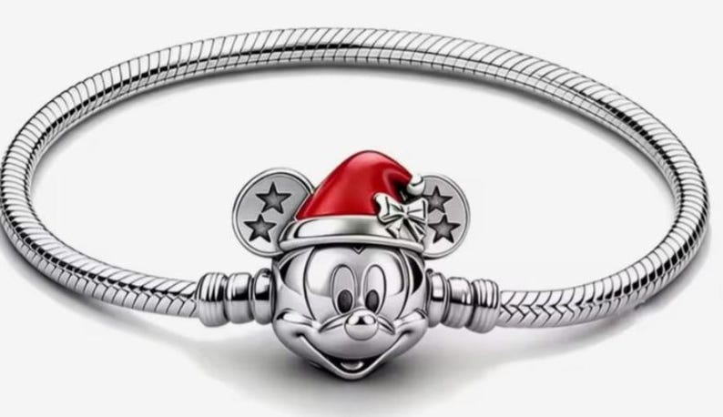 May include: A silver bracelet featuring a Mickey Mouse charm wearing a red Santa hat. The charm has Mickey's face and ears with star accents. The bracelet has a snake chain design.