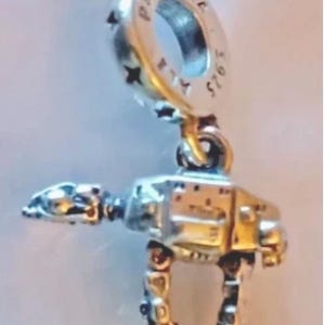 May include: A silver-toned metal charm shaped like a four-legged robotic walker. The top bail is engraved with the text PANDORA and S925. The charm features a detailed mechanical body with articulated legs and a prominent head section, crafted in a polished metallic finish.