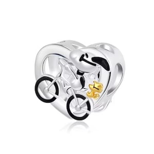 May include: A silver heart-shaped charm bead featuring a polished finish. The design depicts a cartoon dog riding a bicycle with black enamel details on the wheels and ears, accompanied by a small, gold-toned bird figure positioned beside the bike.