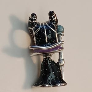 May include: A metal enamel pin featuring a stylized figure with two curved black horns, a black robe with white speckles, a purple collar, and a silver staff topped with a light blue sphere. The figure is depicted from the back view.