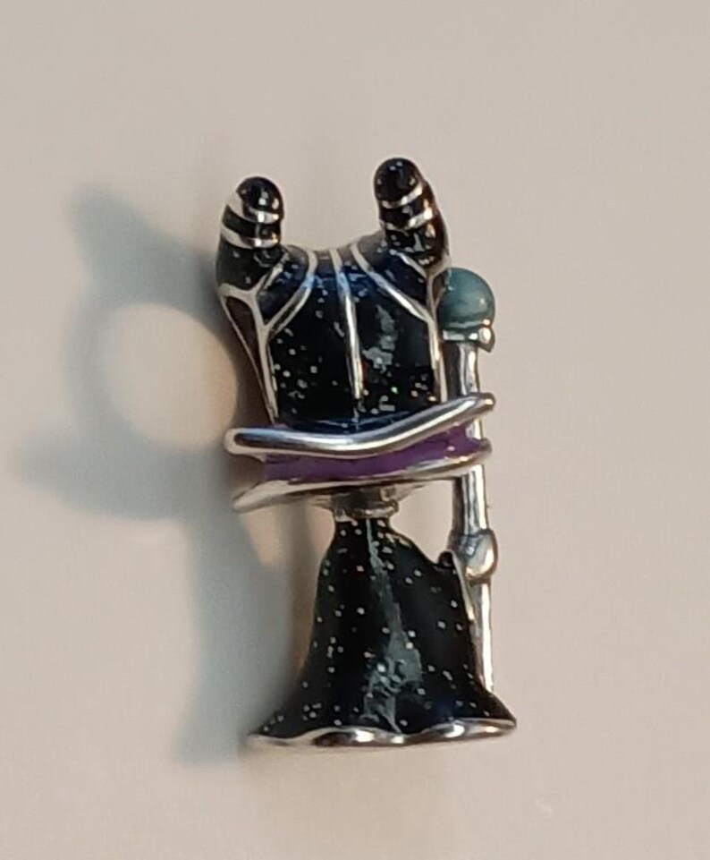 May include: A metal enamel pin featuring a stylized figure with two curved black horns, a black robe with white speckles, a purple collar, and a silver staff topped with a light blue sphere. The figure is depicted from the back view.