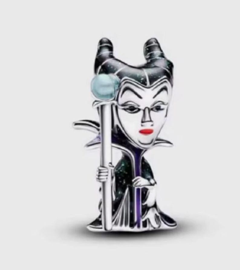 May include: A silver-toned metal charm featuring a stylized character with two prominent horns, a pale face, and bright red lips. The figure wears a dark, glittery robe and holds a long staff topped with a light blue sphere in one hand.