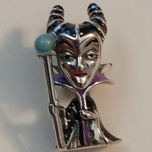 May include: A silver-toned metal pin featuring a character with two curved horns, wearing a black and purple robe with a high collar. The character has a pale face, red lips, and holds a staff topped with a light blue sphere in one hand.