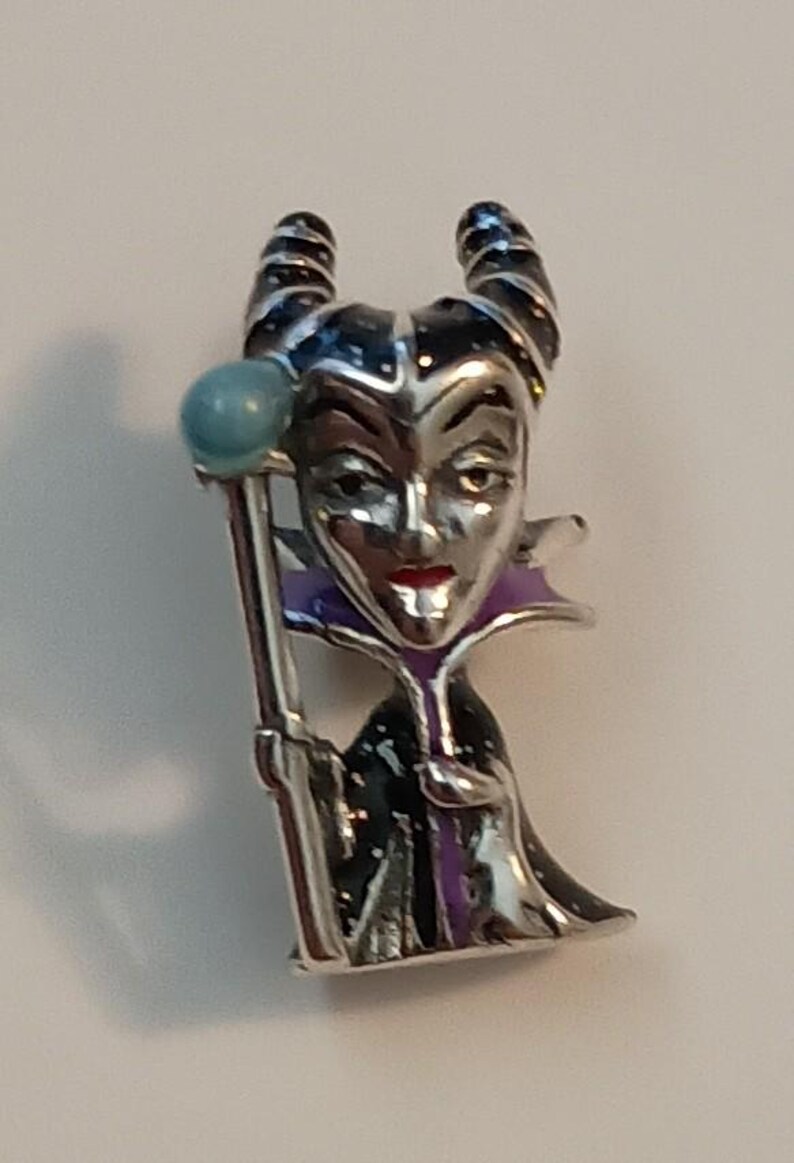 May include: A silver-toned metal pin featuring a character with two curved horns, wearing a black and purple robe with a high collar. The character has a pale face, red lips, and holds a staff topped with a light blue sphere in one hand.