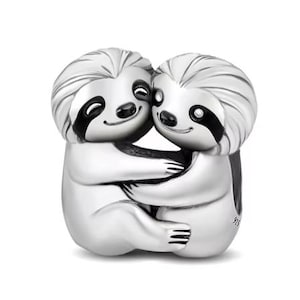 May include: A silver charm featuring two embracing sloths. The sloths have detailed fur, black eyes, and are hugging each other. The charm is made of a shiny metal and is set against a white background. The charm is a cute and whimsical design.