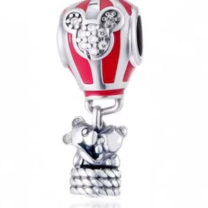 May include: A silver charm shaped like a hot air balloon with red enamel stripes and a Mickey Mouse head silhouette encrusted with clear stones. A silver basket hangs below, featuring two small bear figures and a textured rope-like base detail.