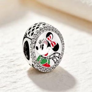 May include: A round silver charm bead featuring a central enamel illustration of a cartoon mouse character with a red bow, a green top, and a black and white checkered scarf, surrounded by a border of small clear crystals set in silver.