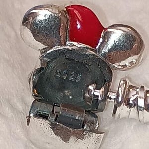 May include: A silver charm with a red enamel accent. The charm has a Mickey Mouse-like design with the text "S925" visible. The charm is attached to a silver bracelet.
