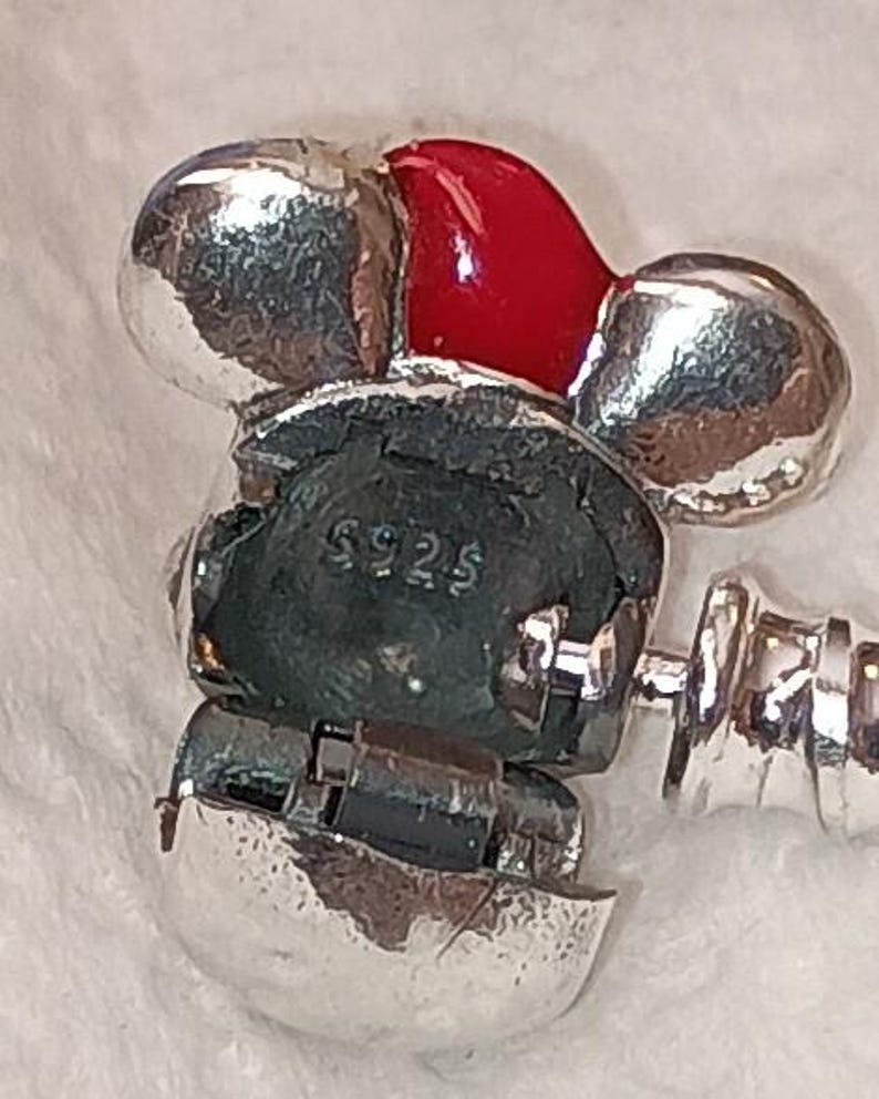 May include: A silver charm with a red enamel accent. The charm has a Mickey Mouse-like design with the text "S925" visible. The charm is attached to a silver bracelet.