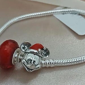 May include: A silver bracelet with a red charm and a Mickey Mouse charm wearing a Santa hat. The bracelet has a snake chain design. The Mickey Mouse charm is silver with a red Santa hat. The red charm is a faceted bead.