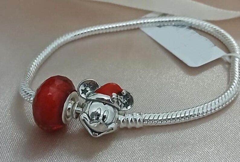 May include: A silver bracelet with a red charm and a Mickey Mouse charm wearing a Santa hat. The bracelet has a snake chain design. The Mickey Mouse charm is silver with a red Santa hat. The red charm is a faceted bead.