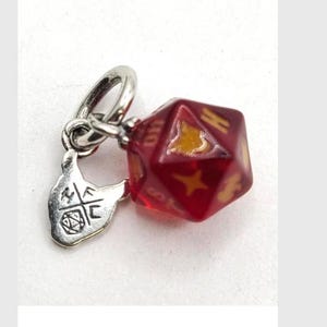 May include: A silver-toned metal charm featuring a translucent red twenty-sided die with gold-colored markings. Attached to the die is a small, flat silver-toned metal tag shaped like a cat head, engraved with the letters HFC and a small die icon.