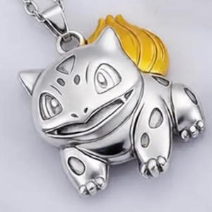 May include: Silver-toned pendant necklace featuring a Bulbasaur design with a gold-colored bulb. The pendant is detailed with visible facial features and body markings, suspended from a silver chain. A charming accessory for fans of the franchise.