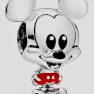 May include: A silver-toned metal charm shaped like a cartoon mouse character with large rounded ears, black eyes, and a black nose. The character is wearing bright red shorts with two white circular accents on the front, rendered in a polished finish.