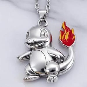May include: A silver-colored pendant necklace featuring a Charmander character design. The pendant has a detailed, three-dimensional form with a red, yellow, and orange flame at the end of its tail. The necklace chain is also silver.