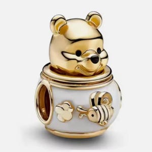 May include: A gold-toned charm bead featuring a sculpted bear head emerging from a white enamel pot. The pot is decorated with a small gold bee and a flower motif. The charm has a central opening for a bracelet, with polished gold metal accents.