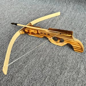 Pode incluir: Besta de madeira com arco castanho claro e corpo castanho escuro. A besta tem uma corda transparente e uma flecha de madeira com ponta preta. O brinquedo de madeira está sobre uma superfície cinzenta.